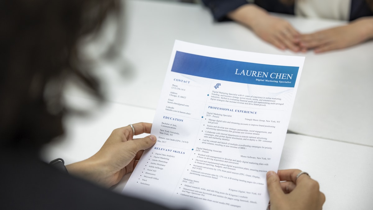 A person reviewing a printed resume at a desk next to an open laptop