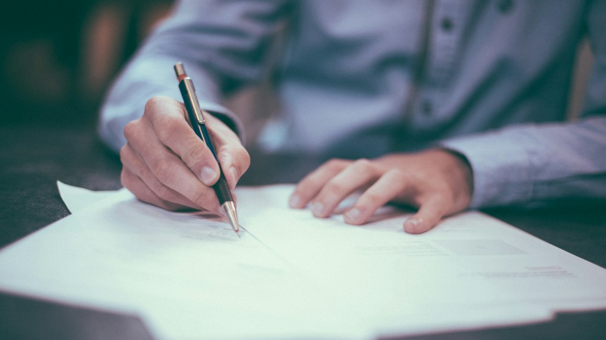 Close-up of a hand signing a printed document at an office desk, pen in hand