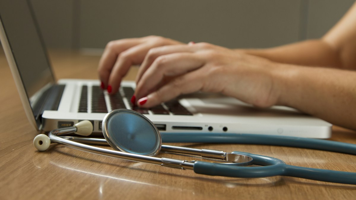 Close-up of hands typing on a laptop keyboard with a stethoscope resting on the desk beside it