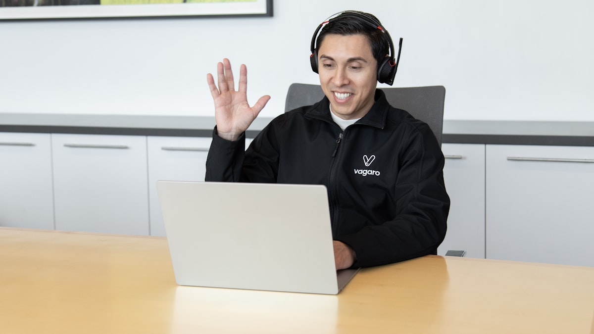 A customer support representative wearing a headset, smiling while assisting a client through a laptop in an office