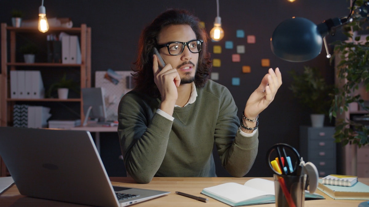 A man at his desk in the evening, smiling while talking on his phone next to an open laptop, representing an after-hours client call