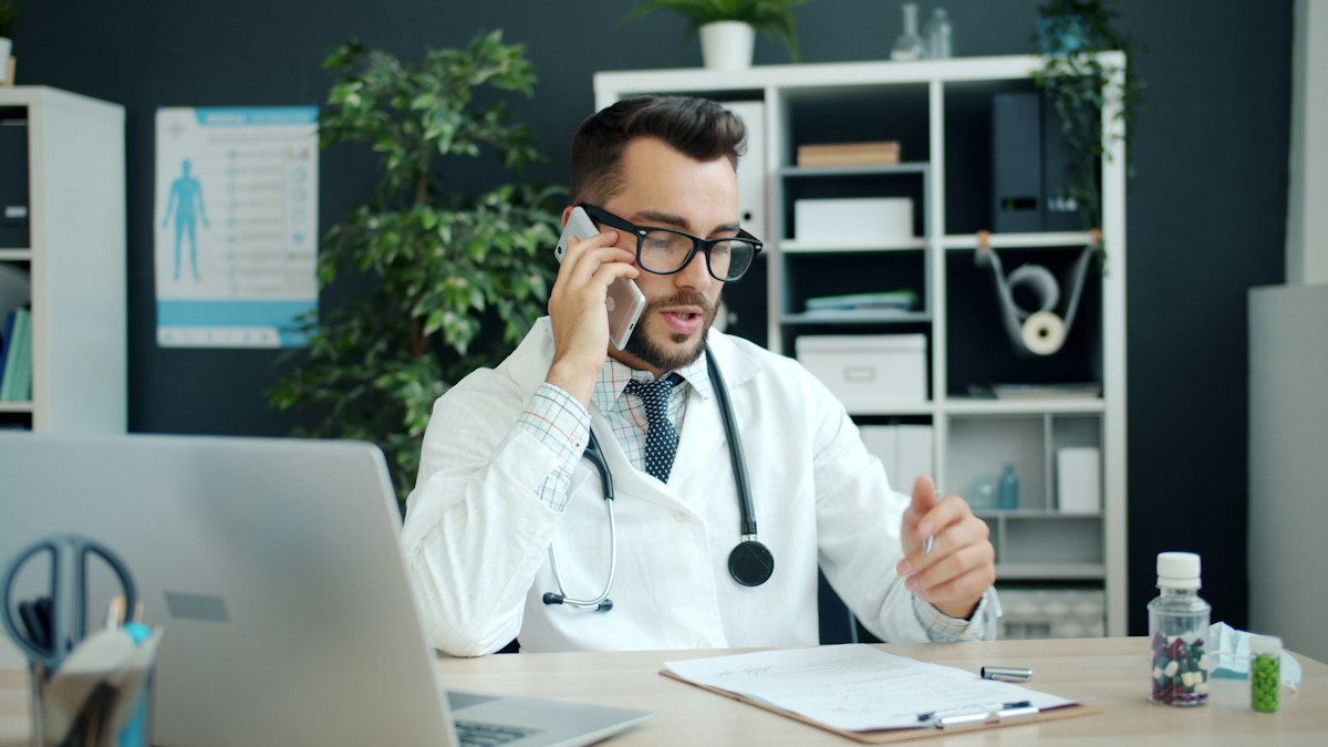 A healthcare professional in a white coat speaking on the phone at a desk while reviewing notes