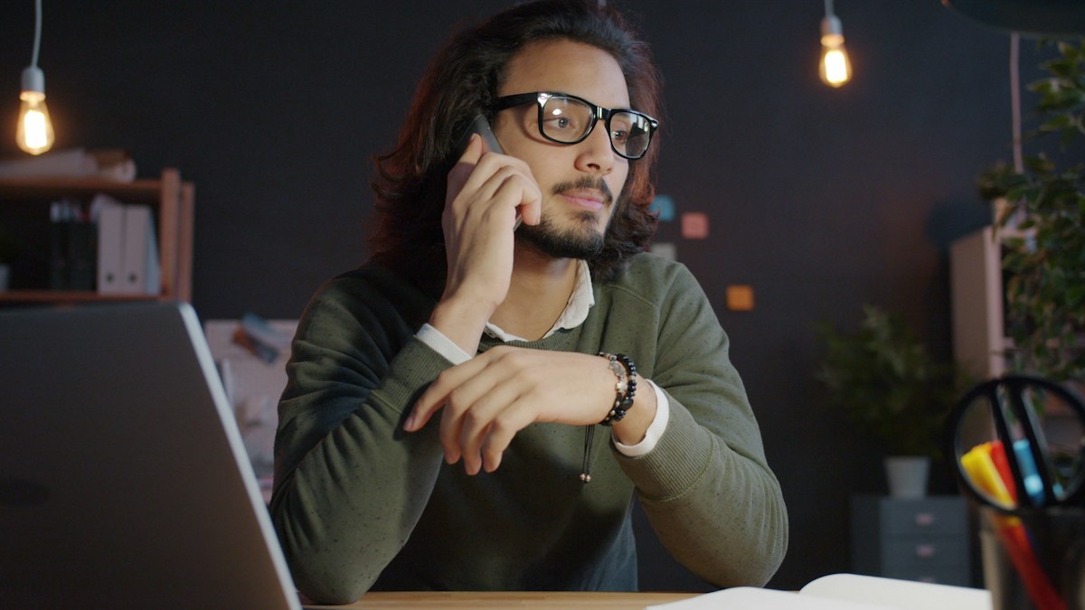 A man speaking quietly on the phone at a desk in a dimly lit office