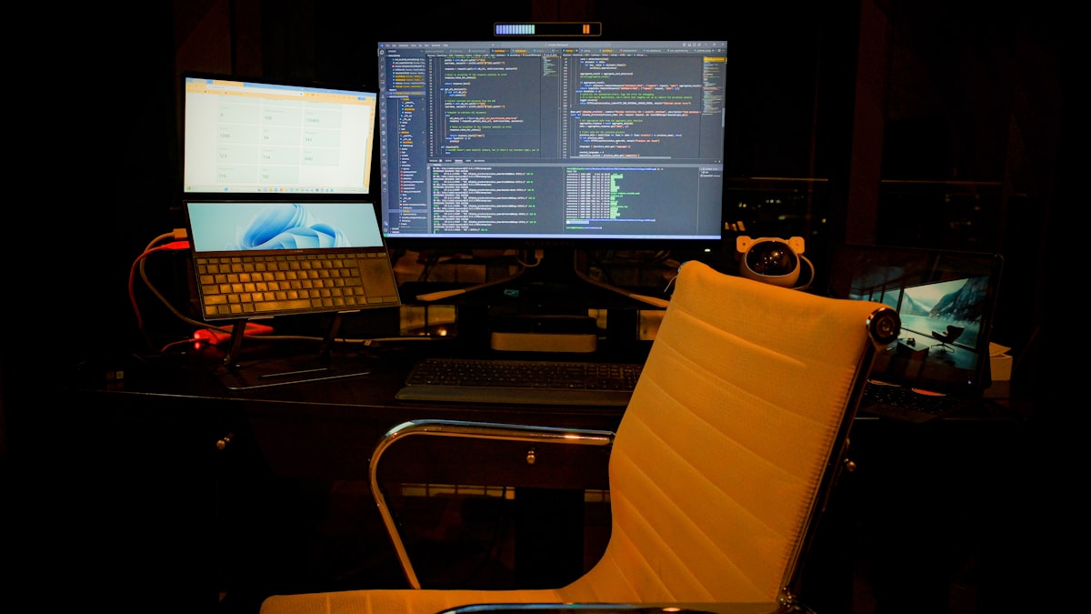 A developer's dual monitor setup showing C# code in an IDE next to a running web application, in an office