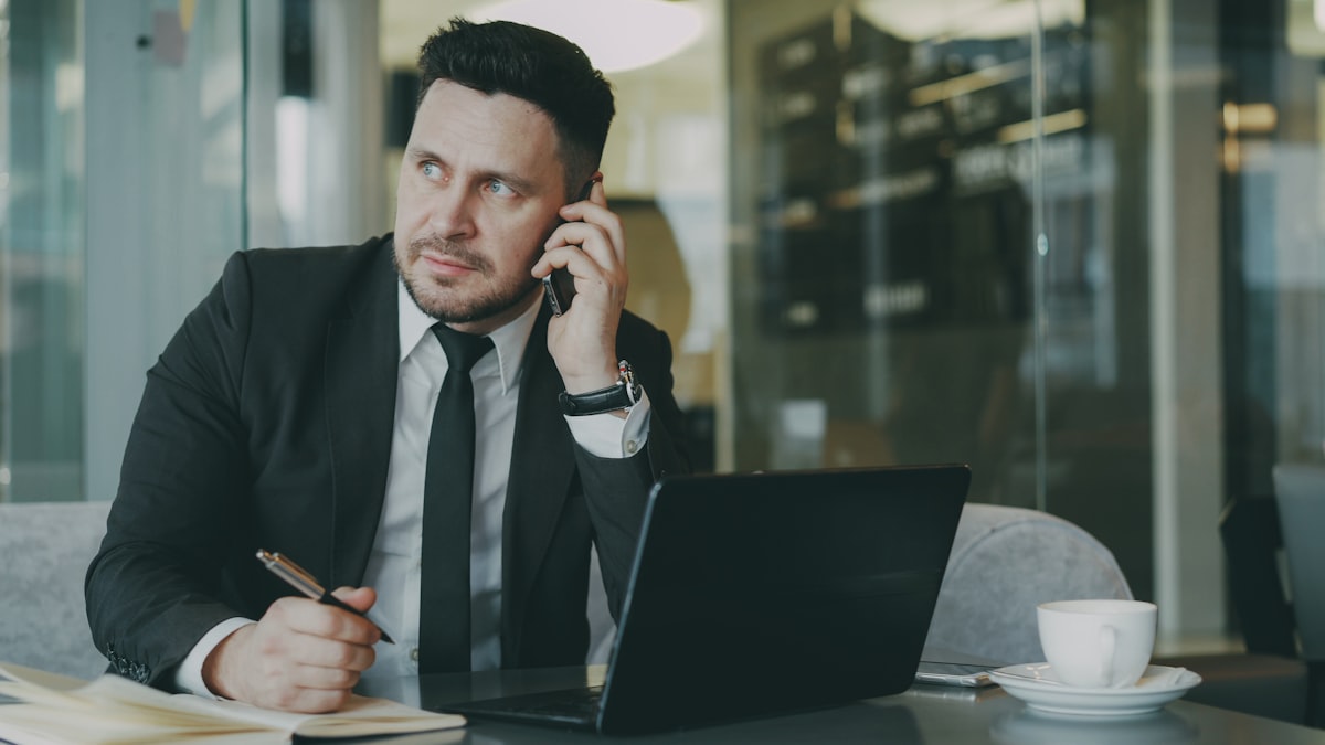 A businessman at a desk talking on his phone while looking at a laptop and writing in a notepad
