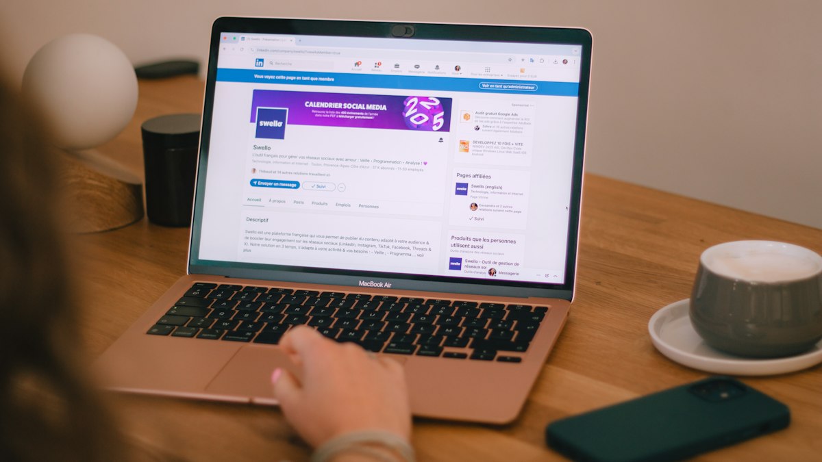 A person's hand on a laptop trackpad with a social media page open on screen, a phone and a cup of coffee on the desk beside it