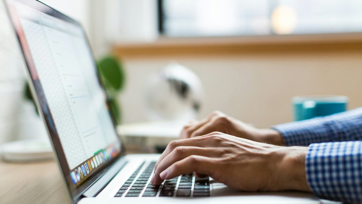 Someone typing on a laptop at a sunlit desk with a plant and a mug nearby, representing a visitor asking a question through a website chat widget